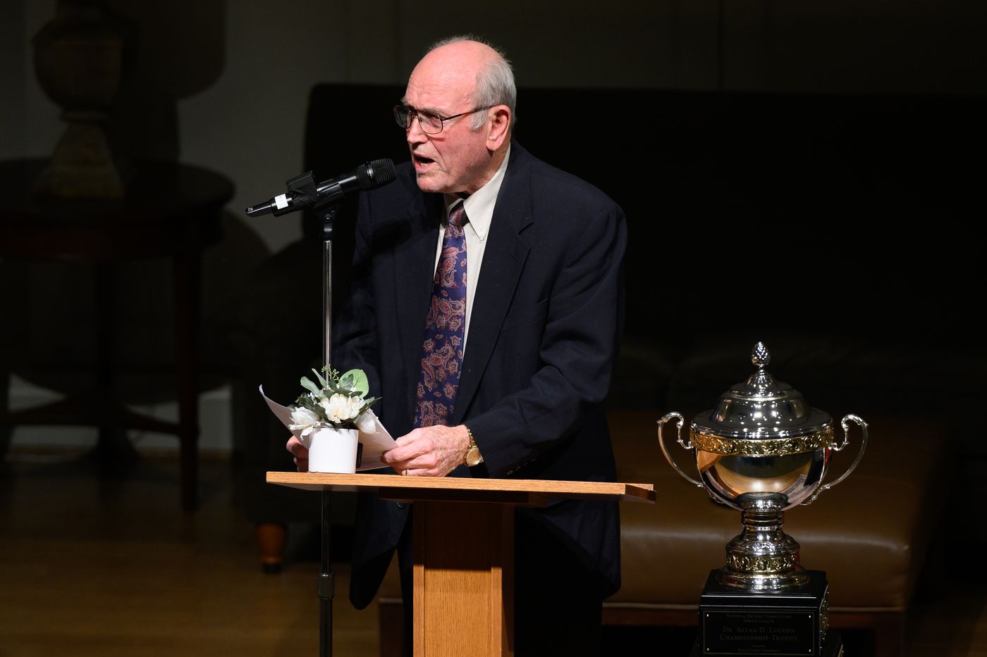 A debater in a dark suit reads from his notes at the podium beside the trophy.