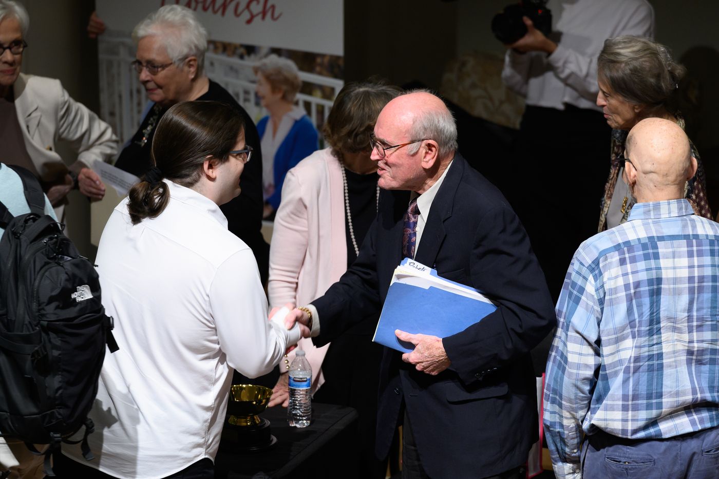 Student coach Cole Wolfe shakes hands with Abbotswood debater Gerald Parker, who holds a blue folder.