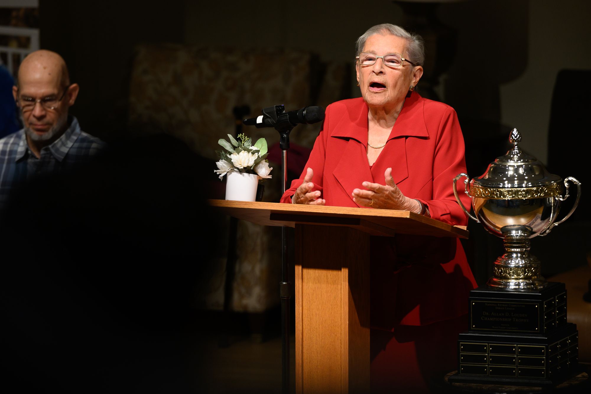 Phyliss Jerome, in a red jacket, speaks at a wooden podium beside the silver championship trophy.